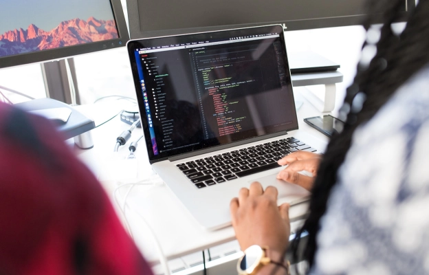 Girl from web development company works in front of the desk with laptop. Behind laptop two monitors present in order to provide web development & design services